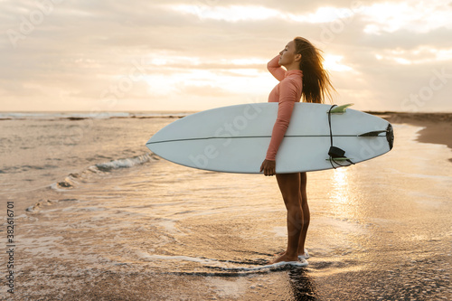 Young attractive surfer woman with white board at sunset on the ocean. Bali Indonesia. Summer time, sports, travel content.