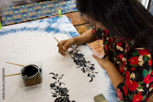 Thai woman people artist painting pattern with natural color indigo on fabric shawl scarf on table in workshop studio at house in Nonthaburi, Thailand