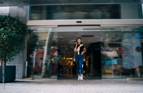 Positive asian hipster girl in trendy wear walking out of building making mobile phone call, smiling beautiful young woman having conversation on smartphone leaving modern business center in town