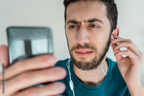 Close up of Man looking at his phone and touching his earphones with awkward face. Concept of broken headphones and hearing problems.