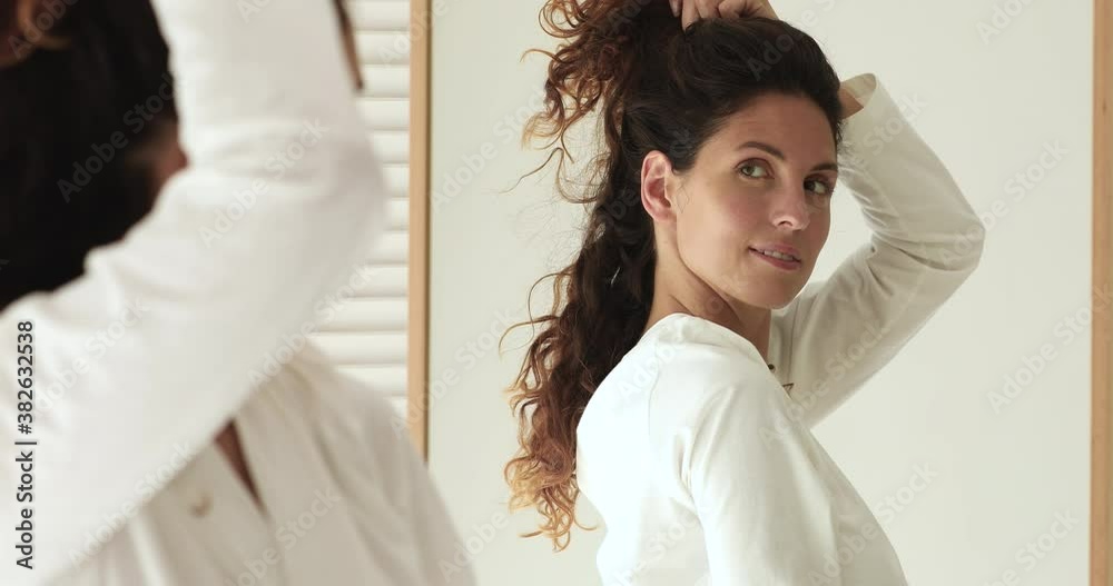 Close up 30s woman in bathrobe freshen up herself in morning looking reflected in mirror touching healthy strong brown curly hairs feels happy. Haircare and styling, best hair products usage concept