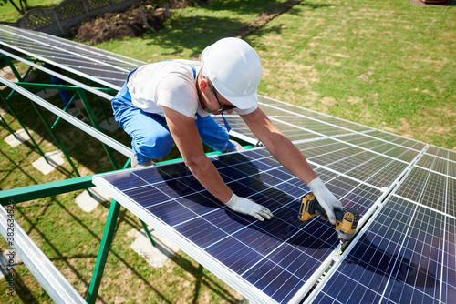 Professional worker installing solar panels on the green metal construction, using different equipment, wearing helmet. Innovative solution for energy solving. Use renewable resources. Green energy.