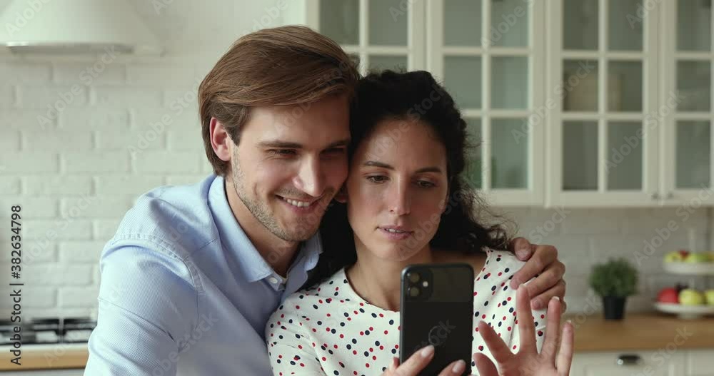 Happy couple using smart phone having fun together at home kitchen, husband hug wife looks at device screen, enjoy new app, scroll feed news watch videos spend time on internet entertaining websites Happy couple using smart phone having fun together at home kitchen, husband hug wife looks at device screen, enjoy new app, scroll feed news watch videos spend time on internet entertaining websites