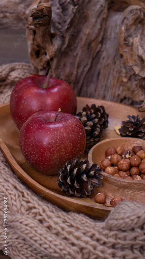 Autumn still life with apples and nuts. Autumn background with apples on a warm knitted scarf, a wooden plate, autumn leaves, hazelnuts and cones.