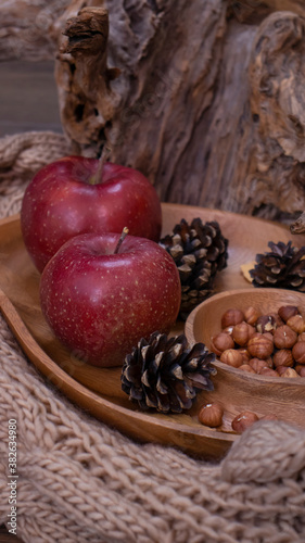Autumn still life with apples and nuts. Autumn background with apples on a warm knitted scarf, a wooden plate, autumn leaves, hazelnuts and cones.