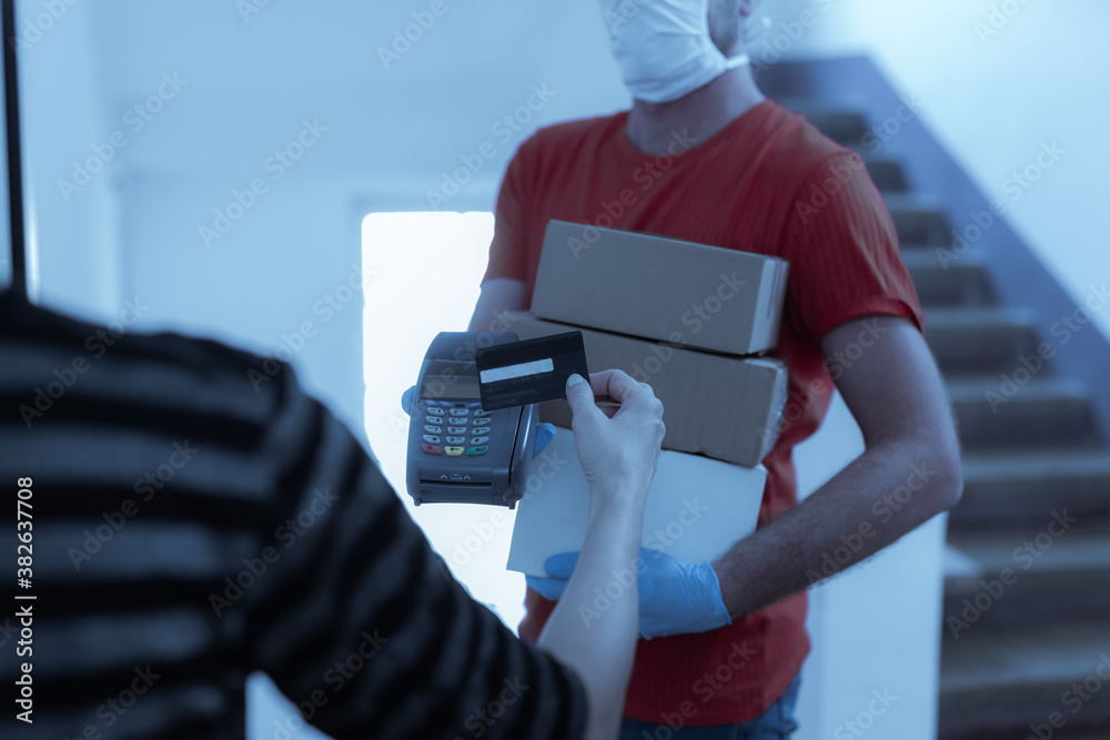 Delivery guy with a medical mask with packages and woman paying with contactless credit card at home during virus pandemic.