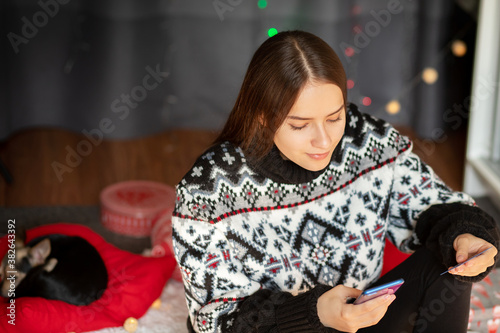 Young girl at home with a small dog chooses Christmas gifts online on the phone from credit card