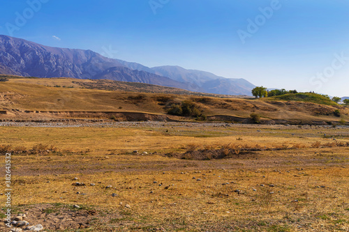 Autumn landscape of the foothills of Kazakhstan. Floodplain of a dried mountain river. Mountains in the background. Blue sky. Foothill vegetation. Dried grass. Herd of horses graze