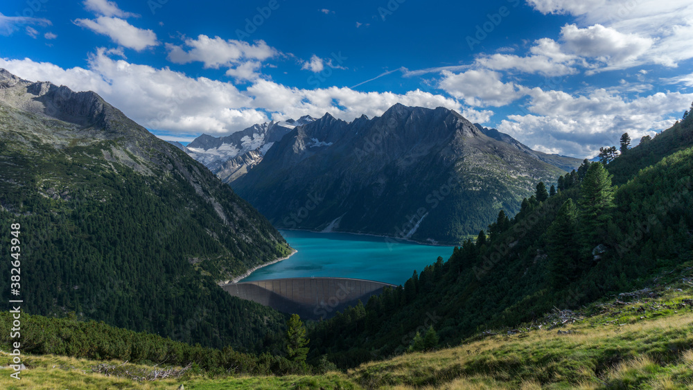 Emerald blue water dam in Alpine mountain range with blue sky and forest