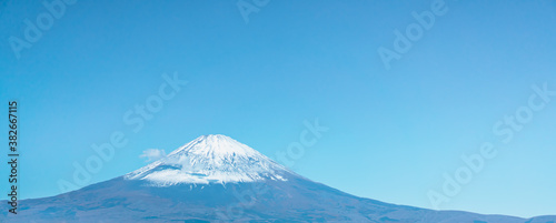 High angle view of Mount Fuji with snow covering the top of the volcano peak on a sunny day of autumn, Japan. There is a bright blue sky with a fluffy white cloud in a soft tone. There is copy space.