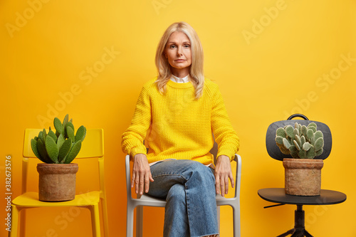 Confident serious female pensioner poses between two chairs with cactus has confident gaze at camera calm expression wears yellow sweater and jeans has rest at home being alone feels melancholic