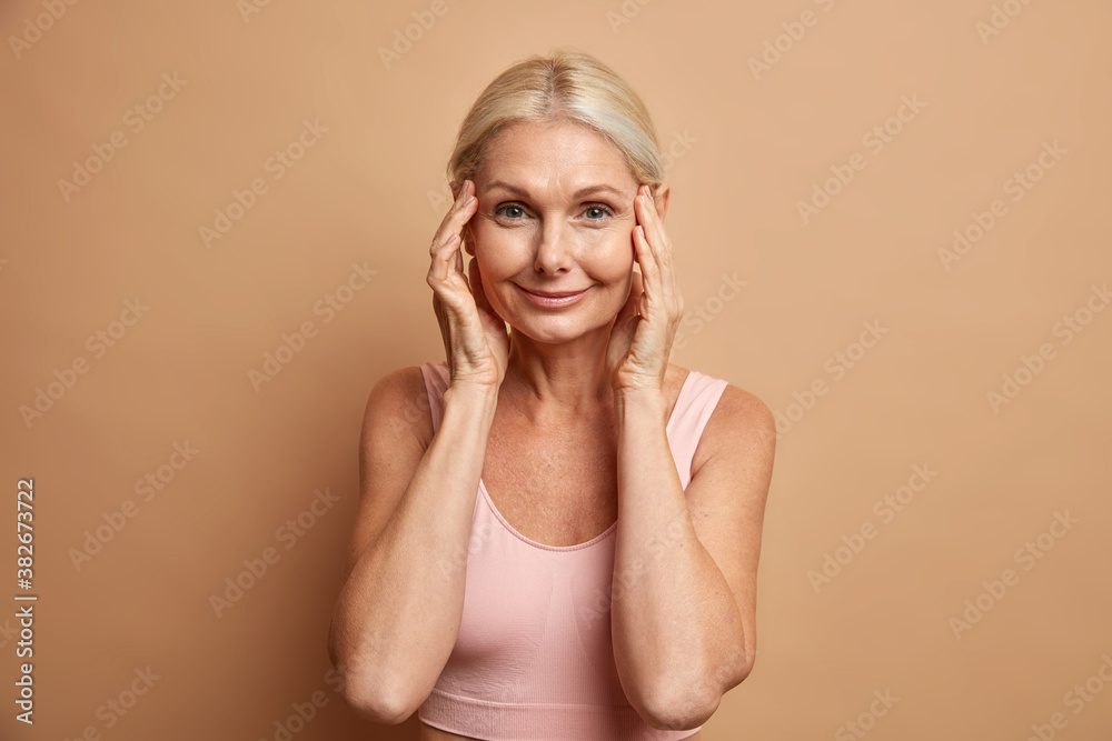 Pretty charming middle aged woman with combed hair touches her perfect skin has minimal makeup well cared complexion takes care of appearance poses in casual top against beige studio background Pretty charming middle aged woman with combed hair touches her perfect skin has minimal makeup well cared complexion takes care of appearance poses in casual top against beige studio background