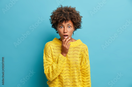 Cute amazed curly haired woman finds out shocking news stares at camera with wide opened mouth stands shocked and astonished wears casual yellow jumper isolated on blue background. Human reaction