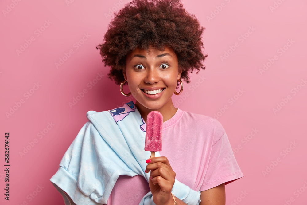 Photo of happy surprised dark skinned woman with Afro hair holds delicious popsicle and dressed casually enjoys eating tasty ice cream poses against rosy background. Summer time and rest concept