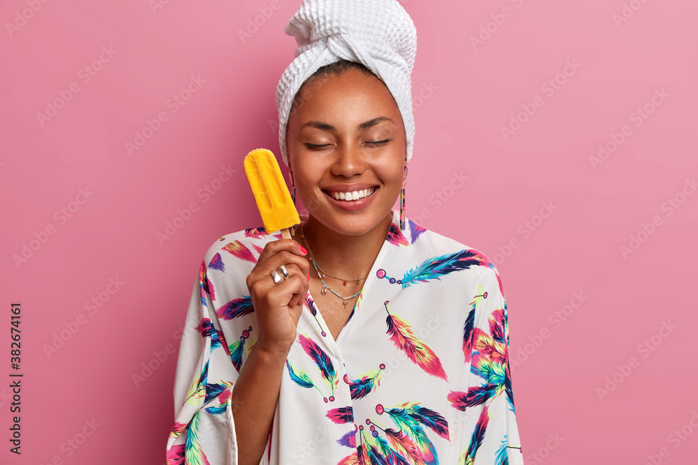 Photo of happy healthy woman with dark skin closes eyes and smiles pleasantly holds delicious ice cream with mango flavor wears domestic robe wrapped towel on head poses over rosy studio wall Photo of happy healthy woman with dark skin closes eyes and smiles pleasantly holds delicious ice cream with mango flavor wears domestic robe wrapped towel on head poses over rosy studio wall