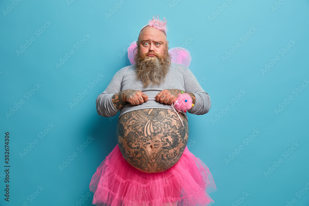 Photo of obese displeased gloomy bearded man fairy with wings holds magic  wand and looks unhappily at camera organizes costume party or holiday  events for children poses against blue background Stock Photo