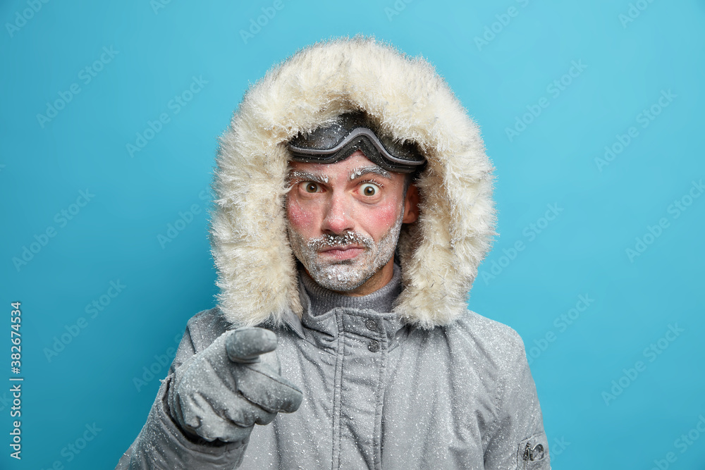 Displeased frozen man with red face points at camera in dissatisfaction blames you wears outerwear for cold weather prepares for winter travel or adventure has active rest isolated on blue background Displeased frozen man with red face points at camera in dissatisfaction blames you wears outerwear for cold weather prepares for winter travel or adventure has active rest isolated on blue background