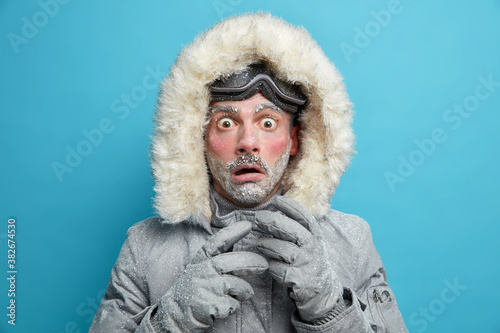 Frozen male explorer has red face covered with frost stares very shocked at camera surprised by very low temperature wears warm jacket and gloves has walk outdoor during blizzard cold weather photo