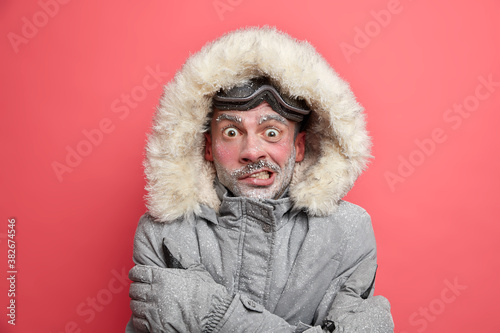 фотографія Frozen man trembles from cold has red face covered by ice frosted beard wears jacket with hood needs to warm during winter expedition poses over coral background