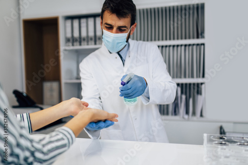 Young male practitioner with face protective mask working at clinic reception desk. He is holding disinfectant bottle and helping his patient to disinfect hands.