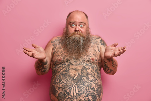 Hesitant fatso man spreads palms in clueless gesture has thick beard has shirtless fat tattoed body poses against rosy background. Unaware bald fatty European guy has no idea feels confused.