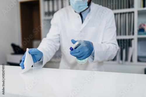 Young male practitioner with face protective mask working at clinic reception desk. He is holding disinfectant bottle and disinfect desk surface. photo