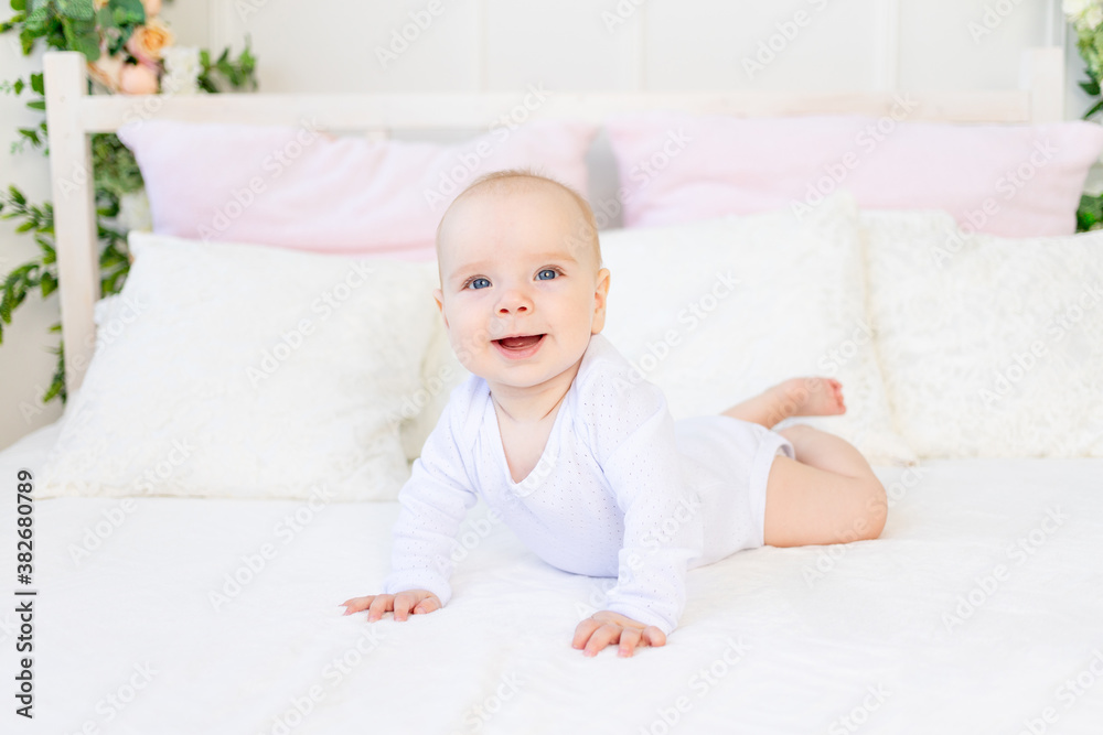 happy baby girl 6 months old in a white bodysuit lying on her stomach on a white bed at home happy baby girl 6 months old in a white bodysuit lying on her stomach on a white bed at home