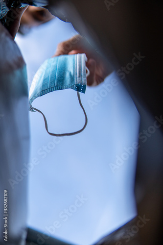 A hand throwing a medical face mask/cloth (3 layers) to a trash bag with a blue sky above. Protection against COVID-19 pandemic (coronavirus) with selective focus on the surgical mask. View from below
