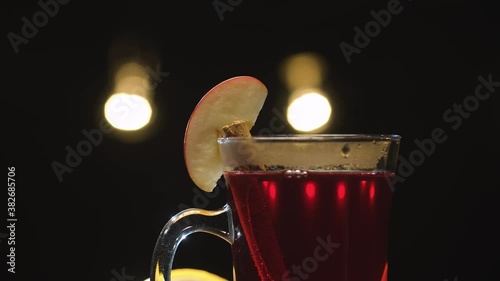 Macro shot hot red mulled wine in a glass with slice of apple and cinnamon stick. Blurry lights on black background. Slide or move camera, dolly shot