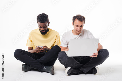 Two mixed race men with phone and laptop sitting on the floor isolated on white background