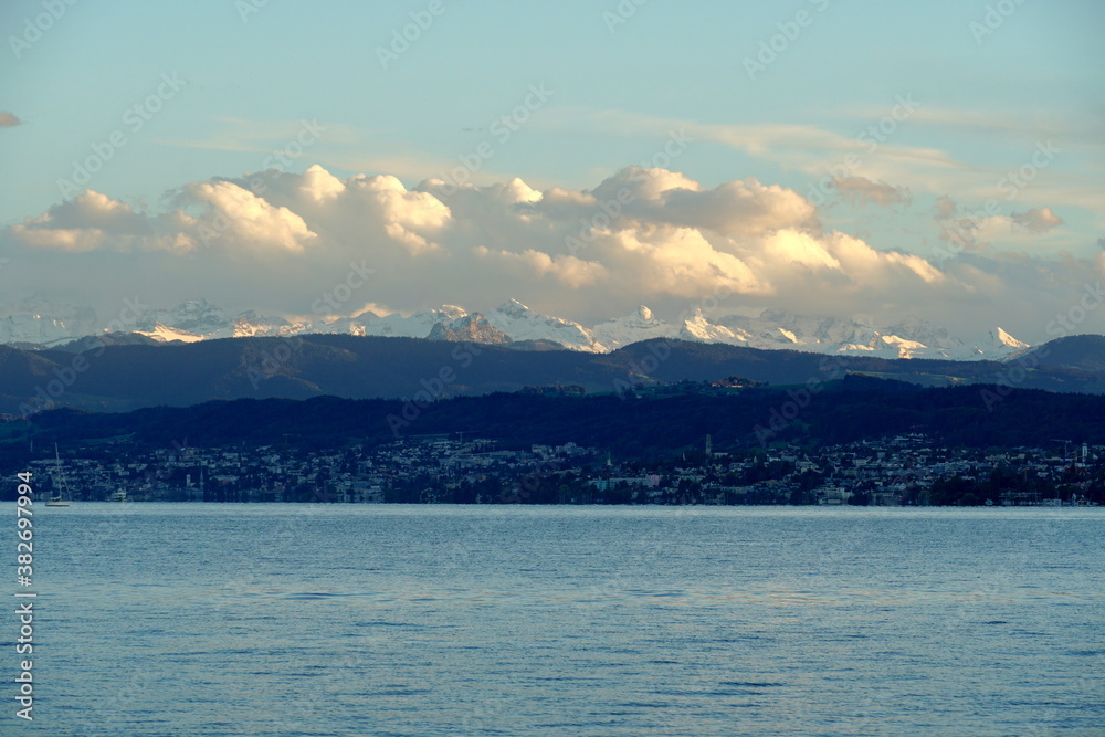 Landscape of the right bank of Lake Zurich with snowy peaks of Alps on the horzizon and cumulus clouds Skyscape enlightened by soft light of sun setting down.