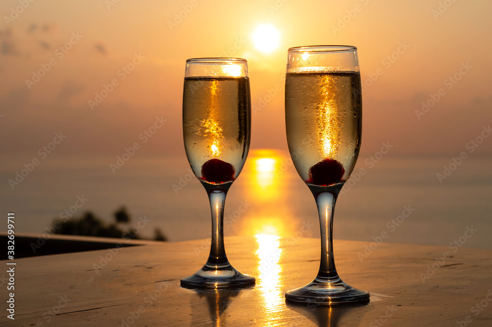 Two wine glasses, with burrowing champagne and cherries, are isolated against the background of the sunset sun on the Mediterranean Sea. Big plan. Romantic background with the ability to insert text. 