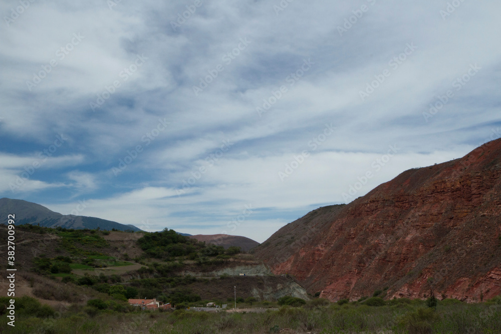 Fototapeta premium Rural scenic. Panorama view of a small farm house, grassland and red sandstone hill under a beautiful sky with clouds.