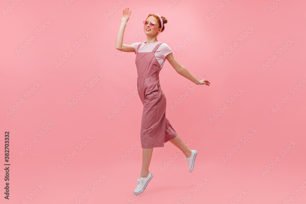 Red-haired woman in overalls and glasses joyfully moves on pink background