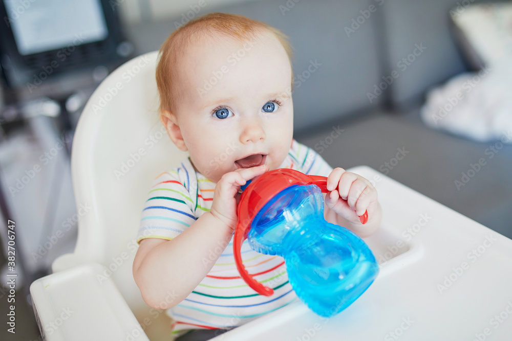 Little baby girl sitting in high chair and drinking water from sippy cup