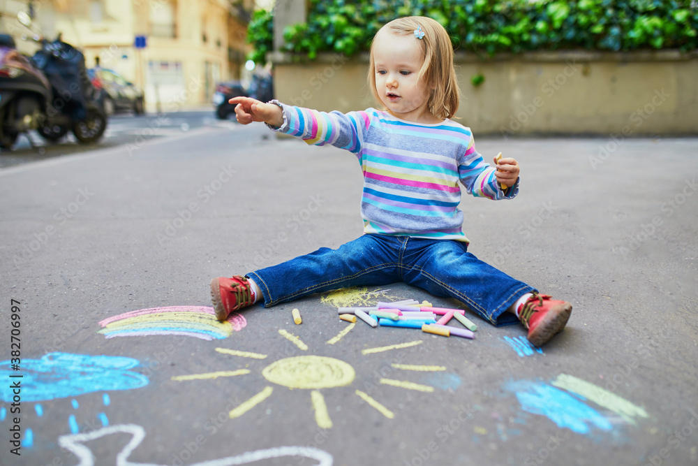 Naklejka premium Adorable toddler girl drawing with colorful chalks on asphalt