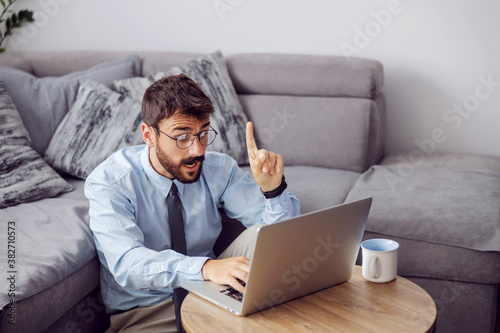 Young attractive bearded businessman sitting on the floor at home, using laptop and having an idea how to solve a problem.