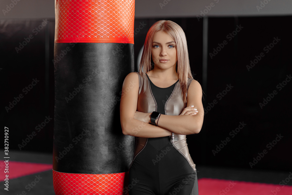 Selective focus of blonde woman in sport wear while standing behind punching bag. Young female fighter posing in empty gym, looking at camera. Concept of sport, boxing.
