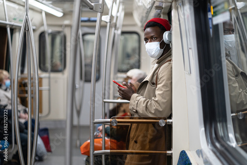 Afro-American passenger man stand in subway train, wear face medical mask to protect yourself from contact with flu virus, covid-19, using mobile phone, listens to music with wireless headphones. 