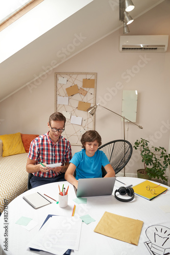 Studying online. Vertical shot of a young smart father wearing eyeglasses helping his little son with school homework, they are watching video lesson on laptop © Kostiantyn