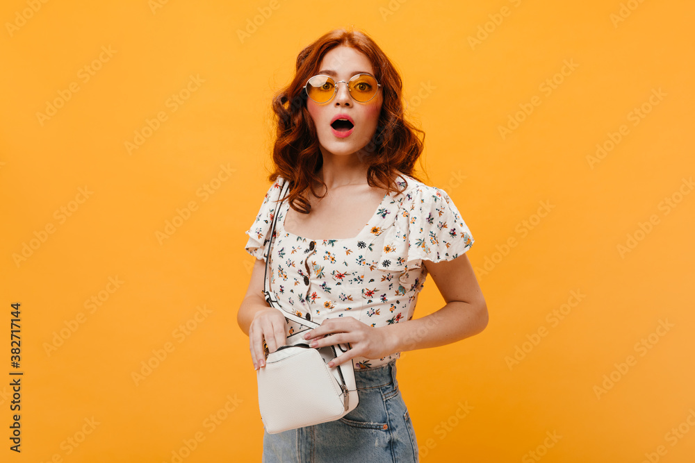 Fototapeta premium Curly girl in sunglasses confusedly looks into camera and holds small white bag
