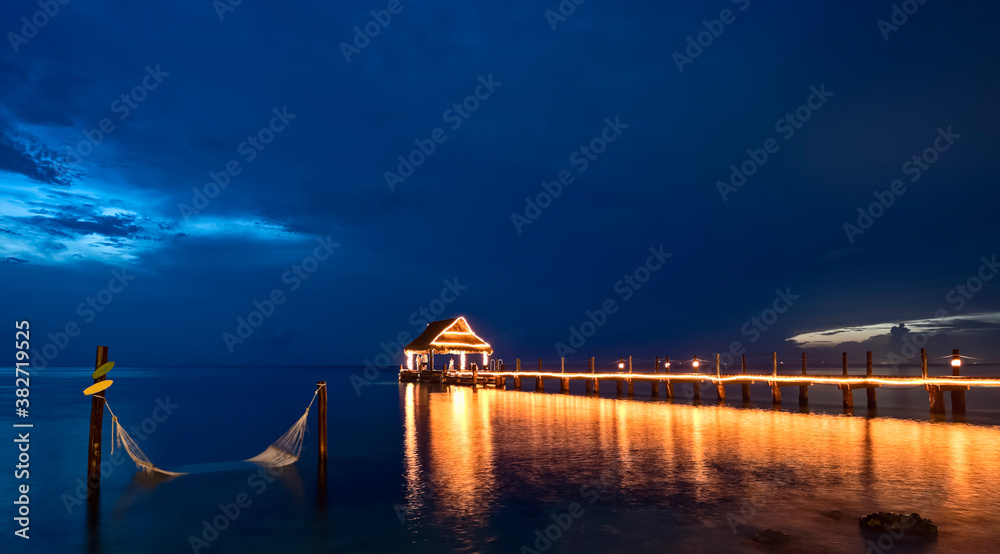 Illuminated wooden fishing pier on the sea after sunset, and a hammock immersed in the Caribbean sea of Cozumel Island, Mexico.