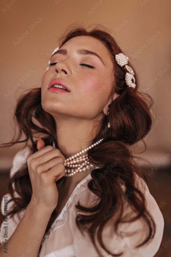 Close-up portrait of woman with freckles holding her pearl necklace. Girl with curly hair posing with closed eyes on beige background