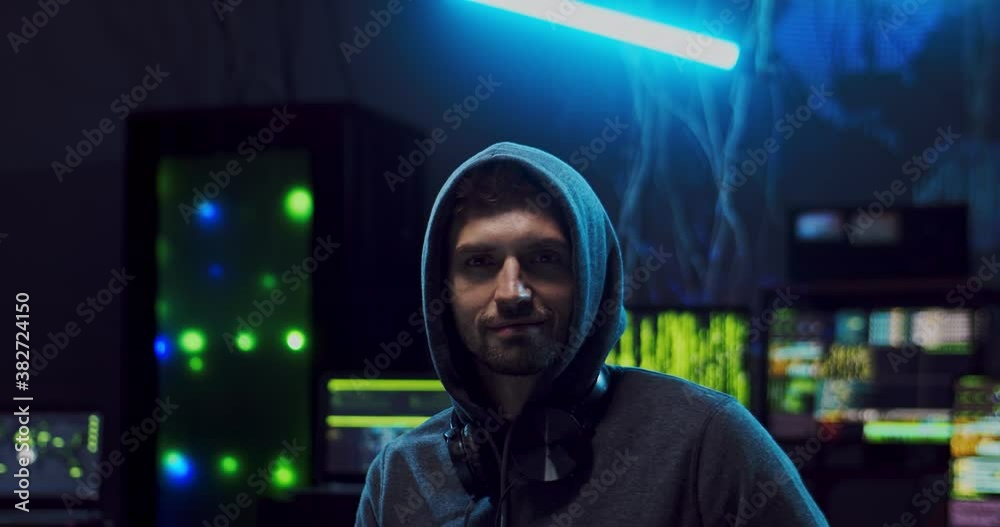 Close up of the young attractive Caucasian guy hacker in a hood looking straight to the camera and smiling cheerfully while sitting in the dark server room with computers on the digital world