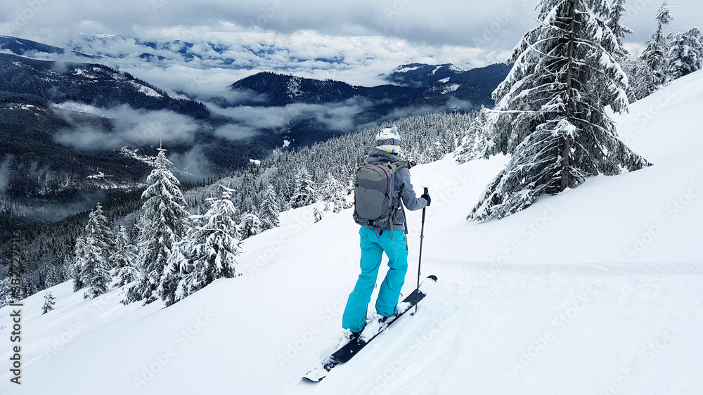 Young skier stands on a gravelly slope with her skis. The mountain slopes are covered with snow. Pine trees covered with snow