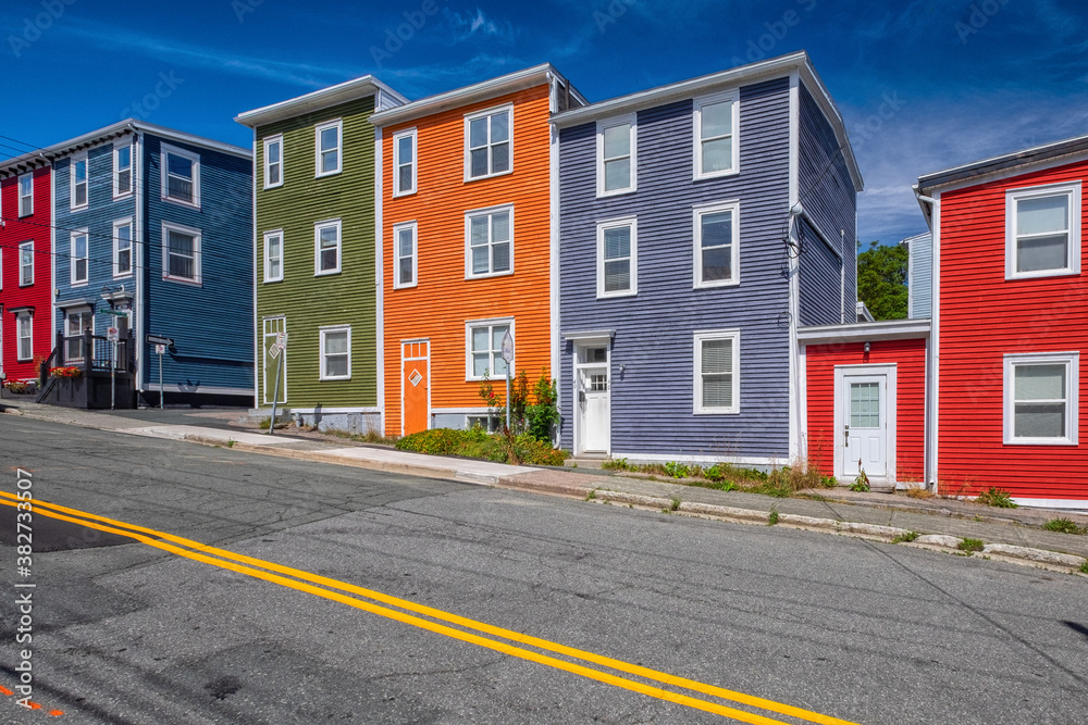 Street view of multiple colorful wooden buildings of various colors. The small structures have double-hung windows and white doors. There's a blue sky in the background.  A street is in the foreground