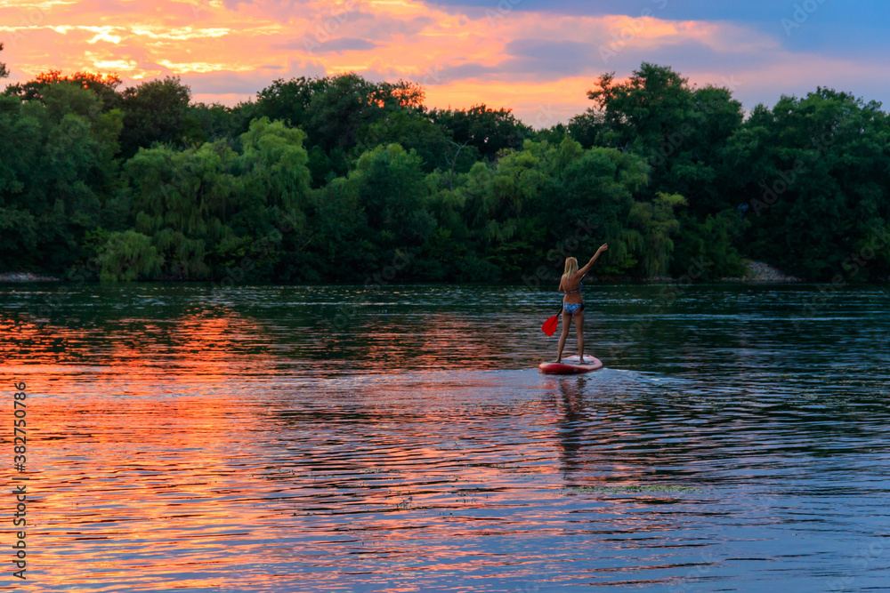 Young woman on stand up paddle board (paddleboard, SUP) paddleboarding along the Dnieper river at sunset