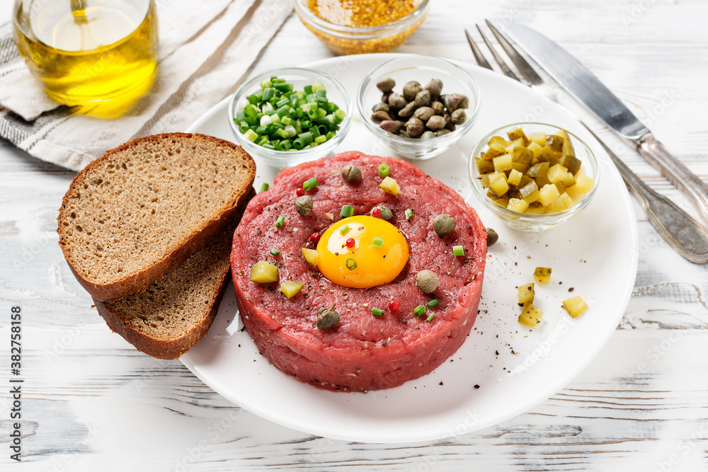 Beef tartare with capers , fresh onion and pickled cucumber on white plate on white wooden background. Beef tartare with capers , fresh onion and pickled cucumber on white plate on white wooden background.