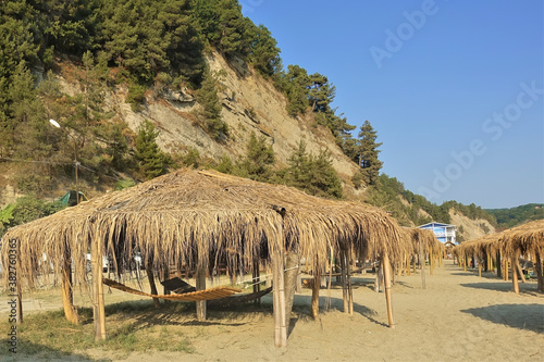 The sandy beach has thatched sheds in a row. Hammocks are suspended below them in the shade. In the background there is a slope of a mountain with coniferous trees. Abkhazia. Pitsunda.
