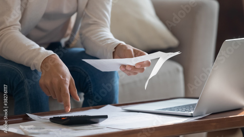 Home office. Close up of young woman owner or renter of dwelling sitting on sofa at desk before laptop screen making calculations of utility payment holding paper bill, invoice or notification in hand